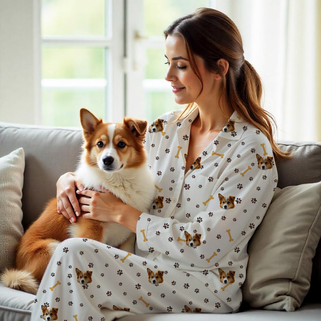 Mujer usando pijama blanco personalizado con la cara de un perro, estampado de huellas y huesos, sosteniendo un perro marrón y blanco en el sofá